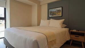 Habitación de hotel con una cama grande, una mesita de noche y un ventanal con vistas a edificios.