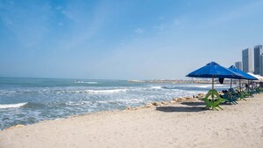 A beach with a row of blue umbrellas and chairs, a rocky shoreline, and a city skyline in the background. - w Ravishing 2BR in the Heart of Bocagrande (Cartagena)