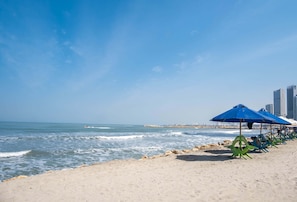 A beach with a row of blue umbrellas and chairs, a rocky shoreline, and a city skyline in the background. - w Ravishing 2BR in the Heart of Bocagrande (Cartagena)