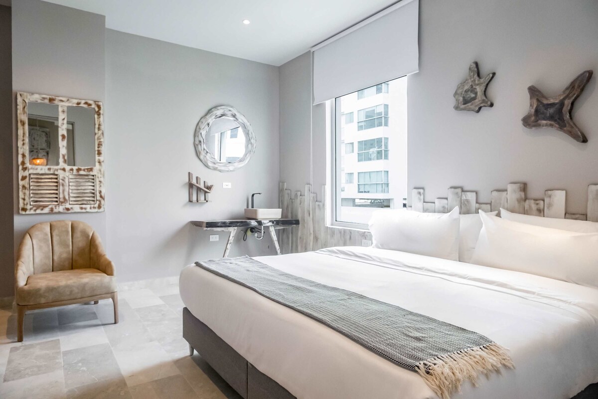 A modern bedroom with a large bed, a chair, a desk, and a window with a city view.