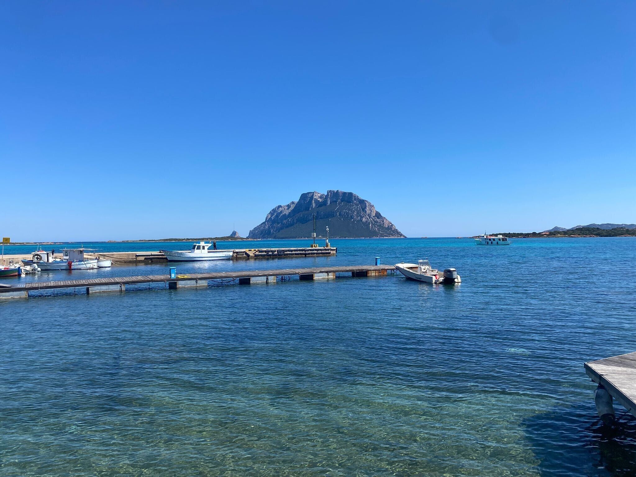 Una marina costera con barcos amarrados, agua azul cristalina y una isla rocosa destacada al fondo.