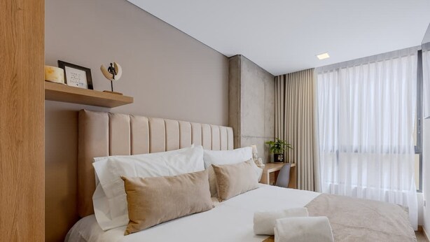 Une chambre d’hôtel moderne avec un grand lit, une tête de lit en bois, une table de chevet avec une lampe et une fenêtre ornée de rideaux légers.