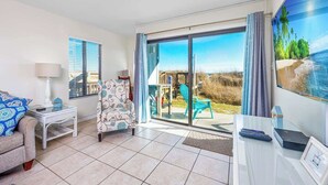 A room with a sofa, a chair, a small table, a TV, and a view of the outdoors through a sliding glass door. - Spacious Beachfront 3br, 2ba Condo in Cherry Grove (Myrtle Beach)