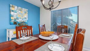 A dining area with a wooden table, chairs, a chandelier, and a large window with blinds. - Spacious Beachfront 3br, 2ba Condo in Cherry Grove (Myrtle Beach)
