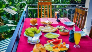 Une table dressée avec une variété de fruits et un verre de jus d’orange, sur un balcon offrant une vue sur une végétation luxuriante.