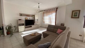 A modern living room with a sofa, ottoman, TV, and a small table with a chess set. - AffittaSardegna - Casa Turris (Porto Torres)