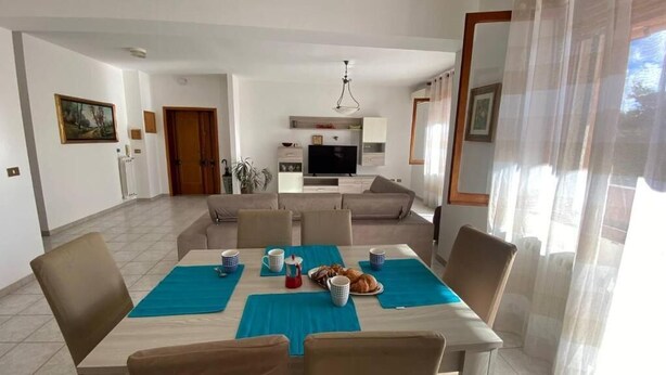 A modern living room with a sofa, a dining table with chairs, and a television in the background. - AffittaSardegna - Casa Turris (Porto Torres)
