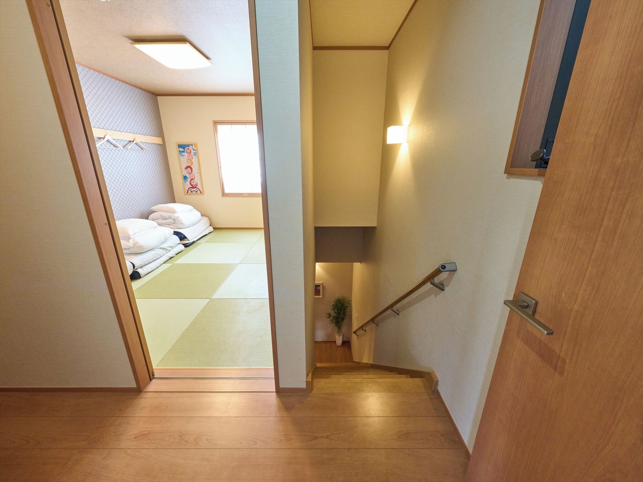 A narrow hallway leading to a bedroom with a bed, a potted plant, and a wooden staircase.