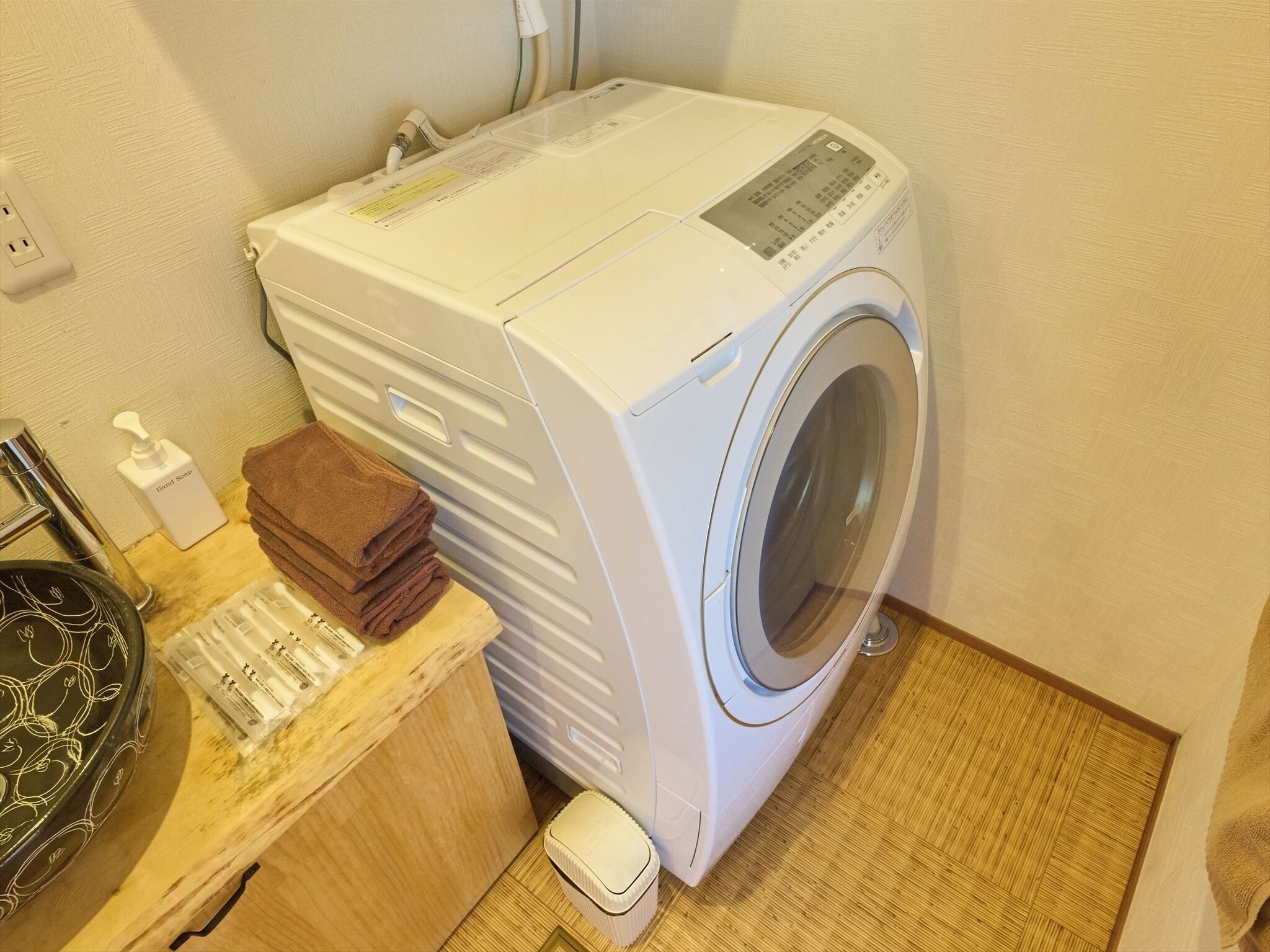 Een compacte, witte wasmachine met voorlader en bedieningspaneel, een klein stapeltje bruine handdoeken en een zeepdispenser op een houten aanrecht.