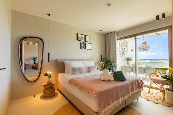 Un dormitorio con cama, espejo, mesita de noche con un florero, balcón con sillas y una mesa, y cuadros en la pared.