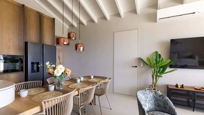 Una cocina moderna con armarios de madera, horno empotrado y refrigerador. Se aprecia un comedor con una mesa redonda, sillas y un florero. En la sala de estar, se observa una planta grande y un televisor.