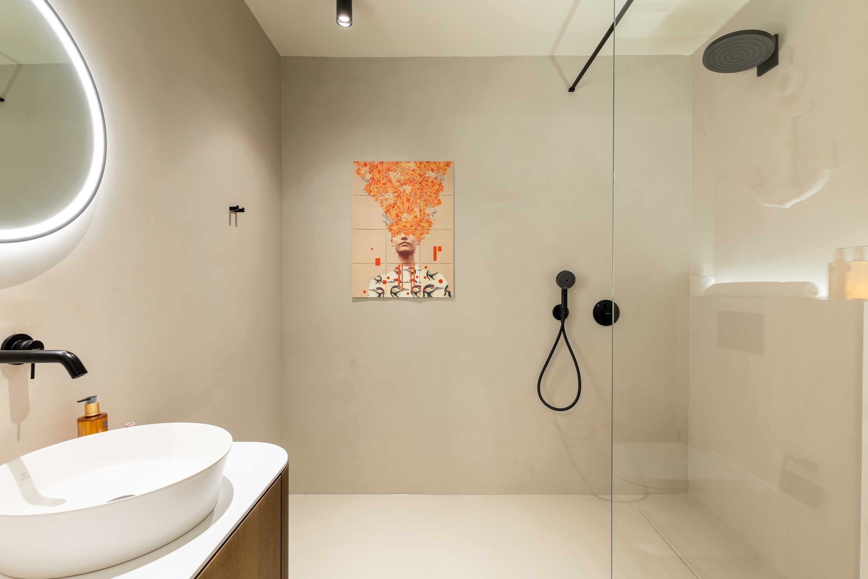 Une salle de bain moderne avec un lavabo blanc, un miroir rond, une douche en verre et une œuvre d’art accrochée au mur.