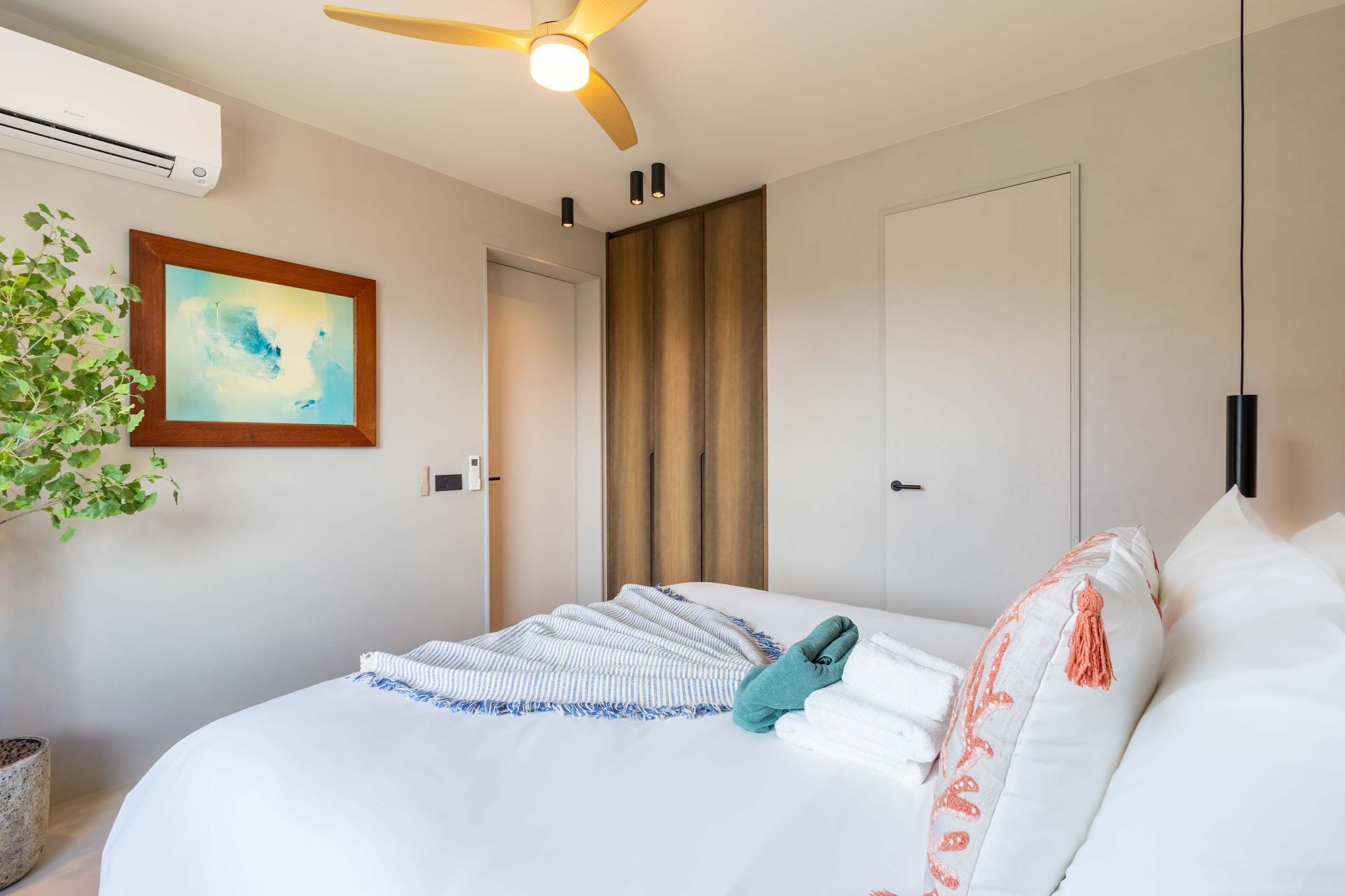 Et hotelværelse med seng, ventilator, aircondition, et indrammet billede, en potteplante og en dør.
