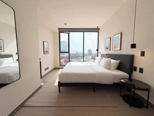 Ein modernes Hotelzimmer mit einem großen Bett, einem kleinen Beistelltisch, einem großen Fenster mit Blick auf die Stadt und einem Spiegel an einer Wand.