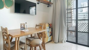 Un salón moderno con una mesa de comedor de madera, sillas, un televisor de pantalla plana, una estantería con plantas y vistas a la ciudad a través de amplios ventanales.