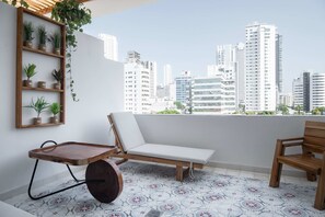 Un balcón con una silla blanca acolchada, un banco de madera y una pequeña mesa de madera con estante para plantas.