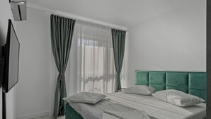 A bedroom with a green upholstered headboard, a bed with white linens, a flat-screen TV mounted on the wall, and sheer curtains. - The Green Oasis (București)