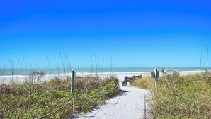 A sandy beach path leading to the ocean, with a bench and a signpost. - New Listing! Beachfront Luxury at Tortuga – Indoor/Outdoor Pool - 3 BDRM (Siesta Key)