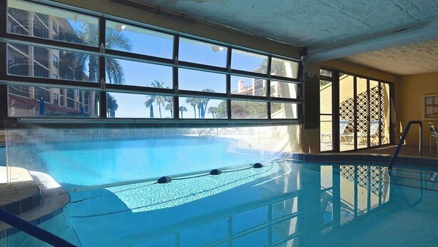 An indoor swimming pool with clear glass walls, a tiled floor, and a view of palm trees and buildings outside. - New Listing! Beachfront Luxury at Tortuga – Indoor/Outdoor Pool - 3 BDRM (Siesta Key)
