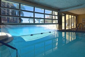 En inomhuspool med väggar av genomskinligt glas, ett kaklat golv och utsikt över palmer och byggnader utanför.