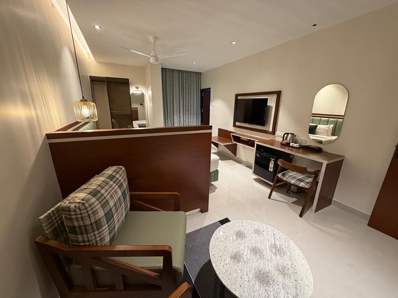 A modern hotel room with a sofa, a round coffee table, a TV, a mirror, and a kitchenette.