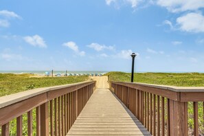 A wooden boardwalk leading to a beach with a lamp post and beach umbrellas in the distance. - 3BR Beachfront Condo w/ Pools & Balcony (South Padre Island)