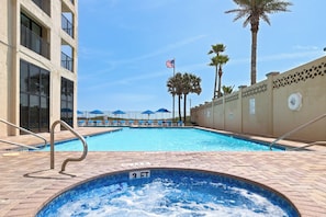 A swimming pool with a 3 FT depth marker, surrounded by lounge chairs and umbrellas, with a palm tree and an American flag in the background. - 3BR Beachfront Condo w/ Pools & Balcony (South Padre Island)