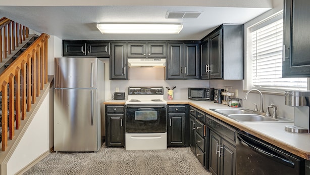 A compact kitchen with dark cabinets, stainless steel appliances, and a wooden staircase.