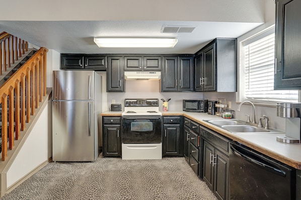 A compact kitchen with dark cabinets, stainless steel appliances, and a wooden staircase. - Walk to Shops: Centrally Located Fort Worth Home (Fort Worth)