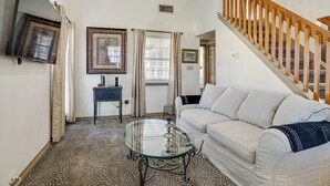 A living room with a glass coffee table, a white sofa, a wooden staircase, and framed artwork on the walls.