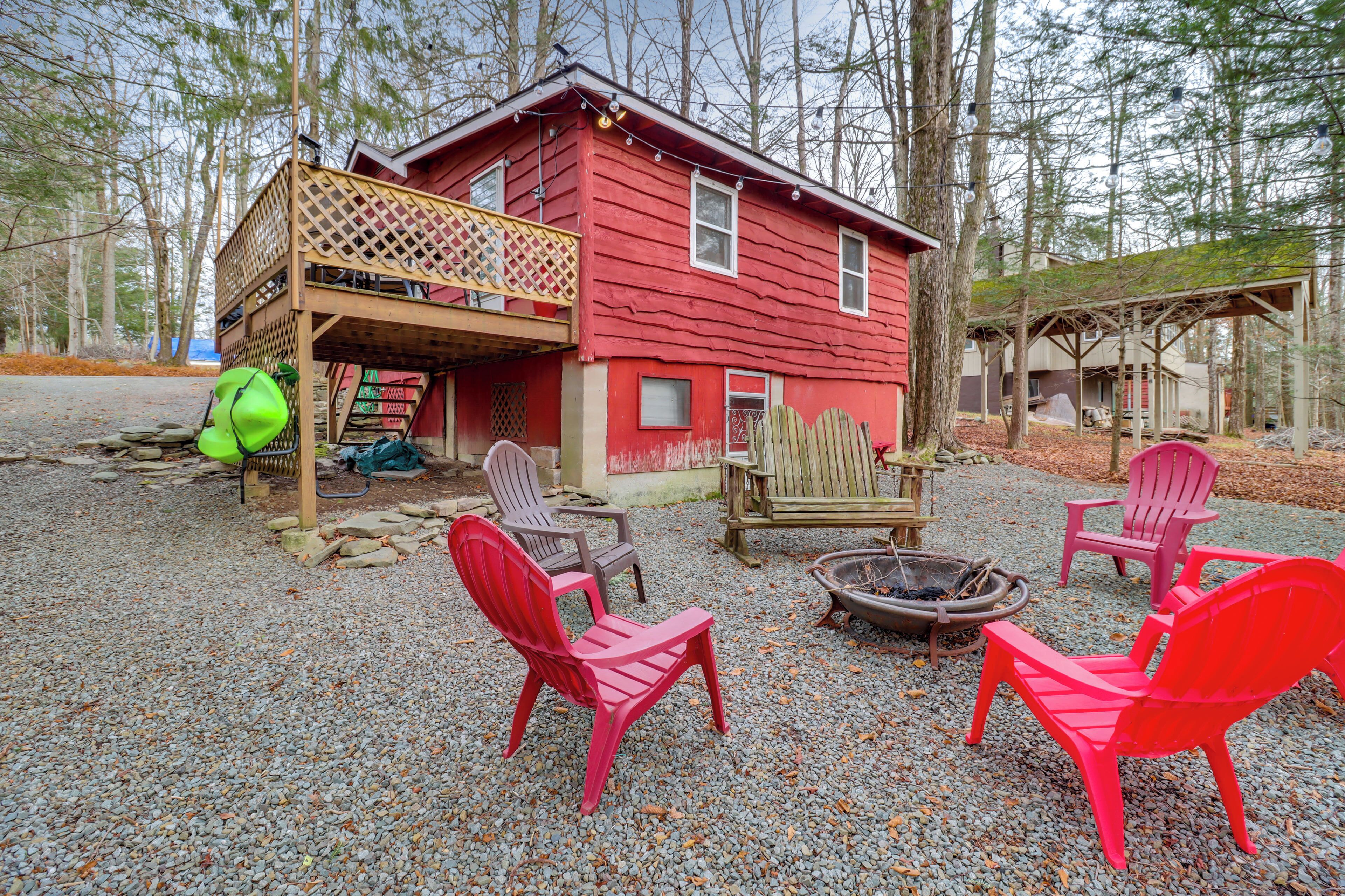 A red cabin with a deck, surrounded by trees. There are four Adirondack chairs and a fire pit.