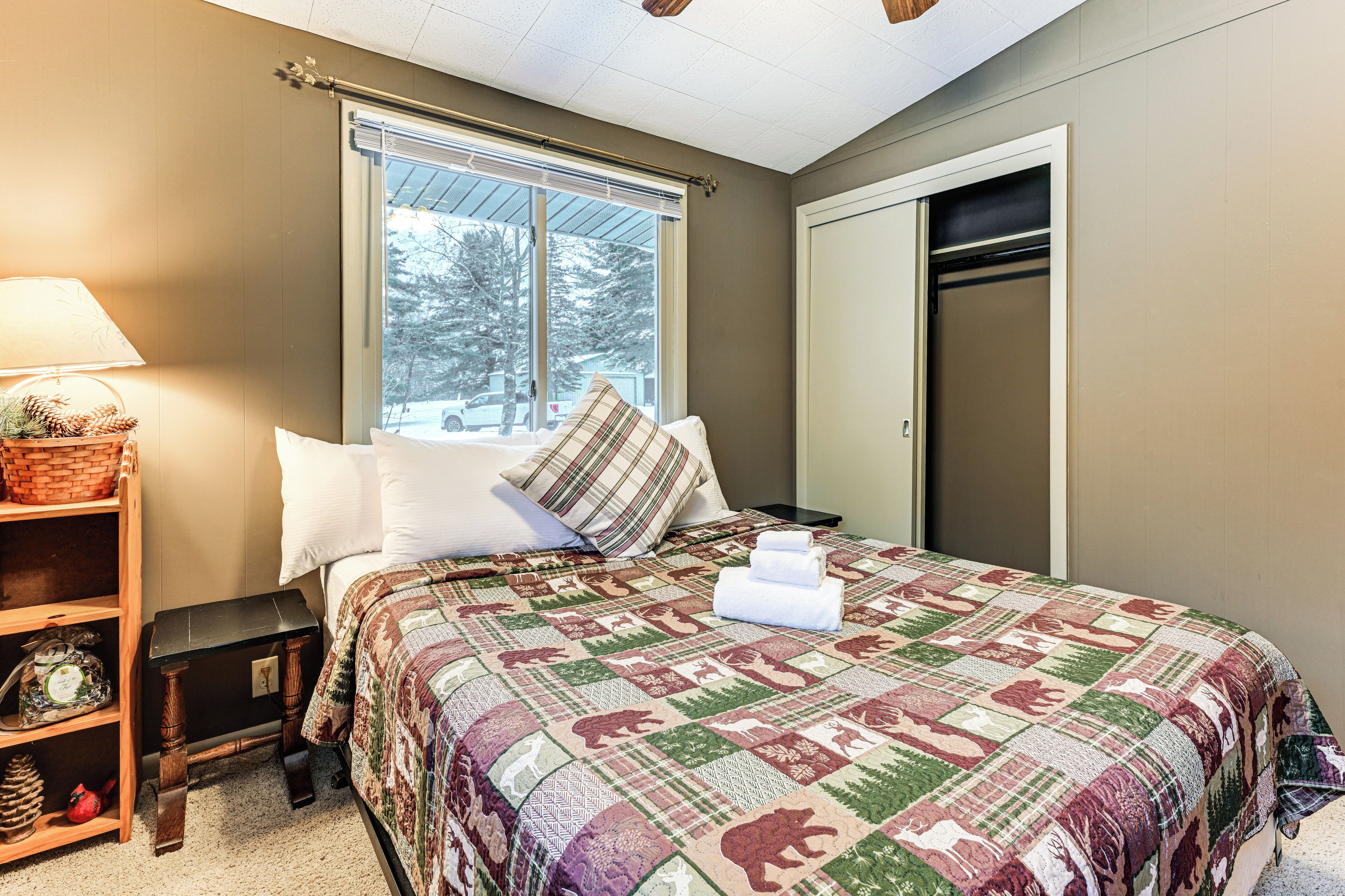 A bedroom with a large bed, a window with a view of trees, a bedside table with a lamp, and a shelf with decorative items.