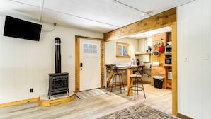 Un salon chaleureux avec une cheminée, un téléviseur fixé au mur et un coin cuisine doté de meubles en bois et d’une fenêtre.