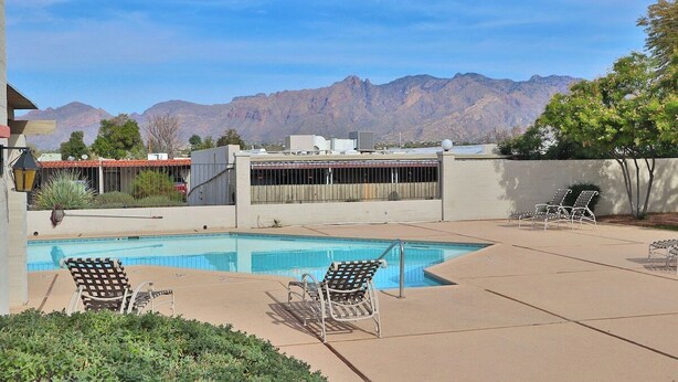 Una piscina con sillones de descanso, montañas al fondo y un edificio con tejado rojo.