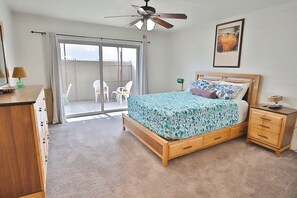 Une chambre à coucher comprenant un lit, une table de chevet en bois, un ventilateur de plafond, une porte-fenêtre coulissante donnant sur un balcon et un tableau encadré accroché au mur.
