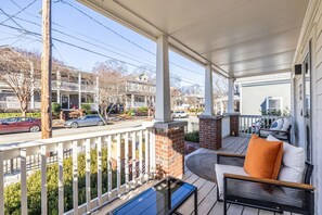 A covered porch with a brick fireplace, a seating area with cushions, and a view of a residential street. - Walk to BeltLine | 3 Miles to World Cup Stadium (Atlanta)