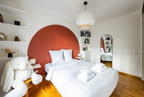 Quarto moderno com cama grande, roupa de cama branca, parede de destaque vermelha, escultura de urso branco e espelho.