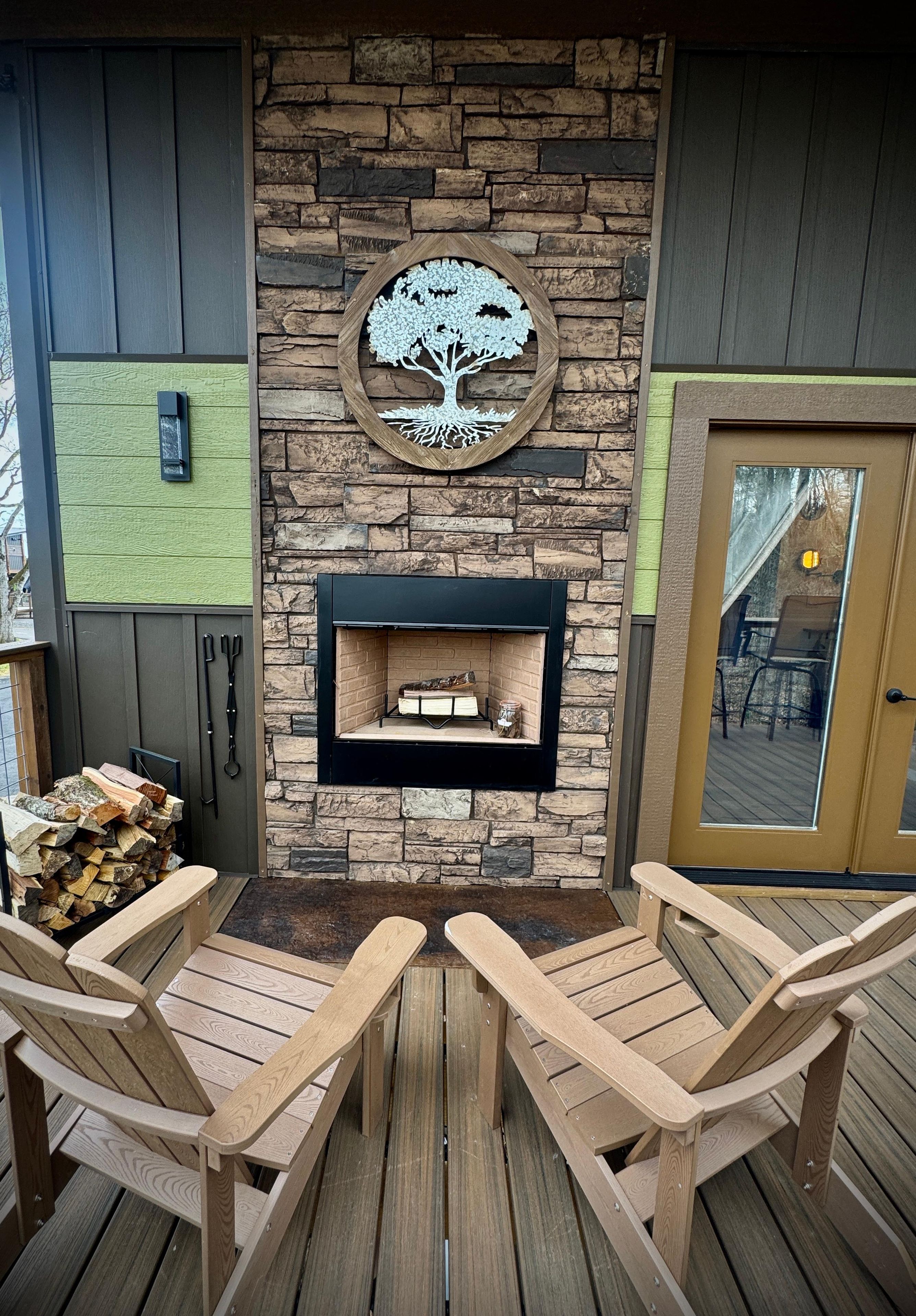 A stone fireplace with a tree design, surrounded by wooden chairs and a firewood stack.