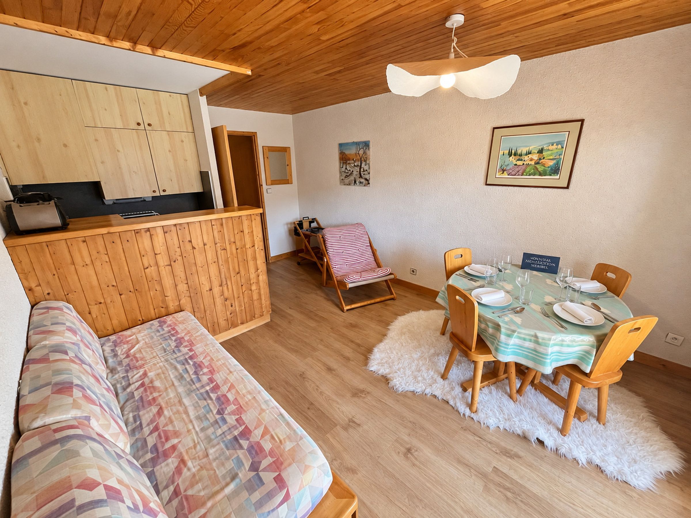 Une pièce chaleureuse avec un plafond en bois, une table à manger dressée pour deux personnes, un canapé confortable et un petit coin cuisine.