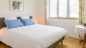 A bedroom with a bed, an air conditioner, a window with curtains, and a small table with a lamp.