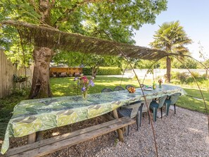 An outdoor dining area with a table covered by a patterned tablecloth, surrounded by chairs, under a shaded canopy.