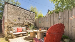 Een houten terras met een rode Adirondack-stoel, een houten bank met kleurrijke kussens en een stenen muur met een klein raam.