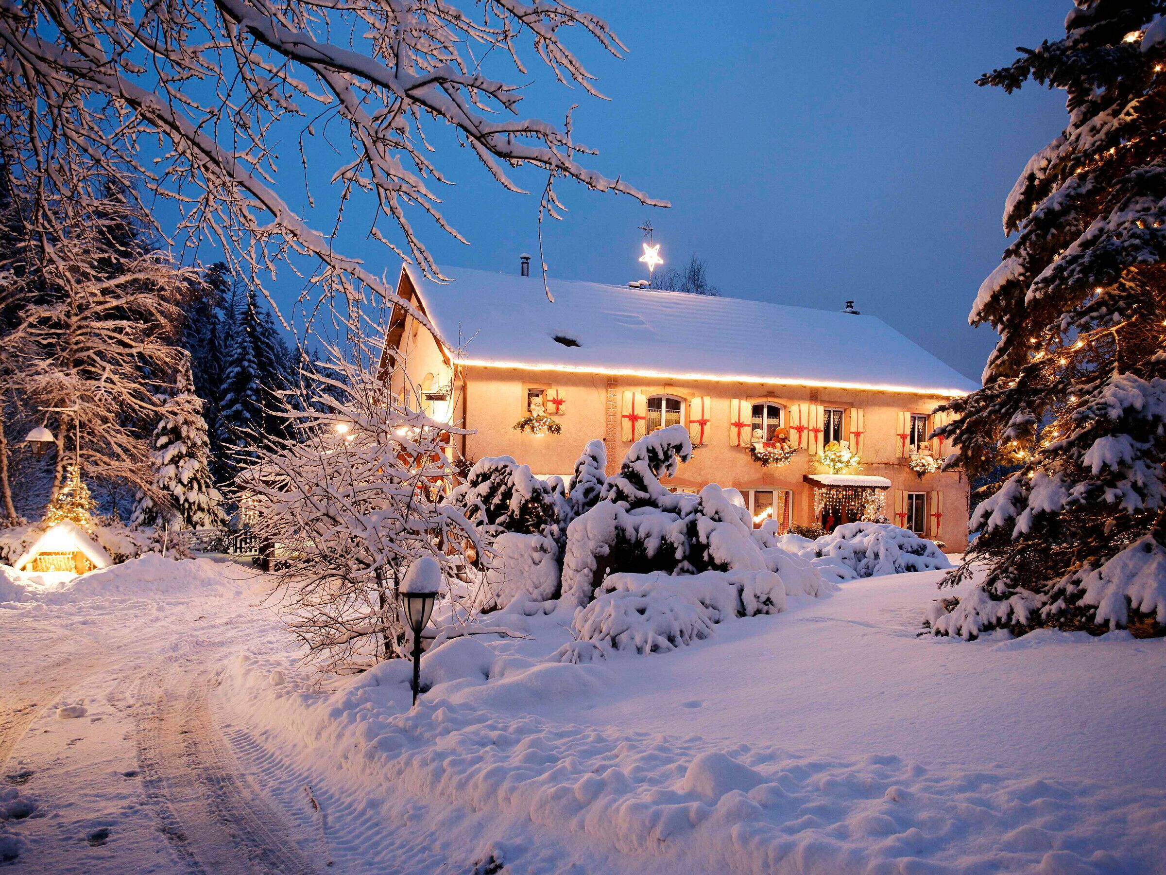 A snow-covered building with a lit entrance, surrounded by snow-laden trees and a snow-covered path.