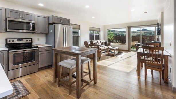 A modern kitchen with stainless steel appliances, a dining area with wooden furniture, and large windows offering a view of the outdoors.