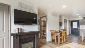 A modern living room with a fireplace, a flat-screen TV mounted above it, a dining area with a wooden table and chairs, and a kitchen area with cabinets and a window.