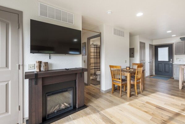 A modern living room with a fireplace, a flat-screen TV mounted above it, a dining area with a wooden table and chairs, and a kitchen area with cabinets and a window.