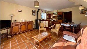 A living room with a sofa, a coffee table, and a TV. There is a kitchen area with wooden cabinets and a dining table with chairs. - 3-room house in Carnac - 4 ppl, terrace and garden (Carnac)