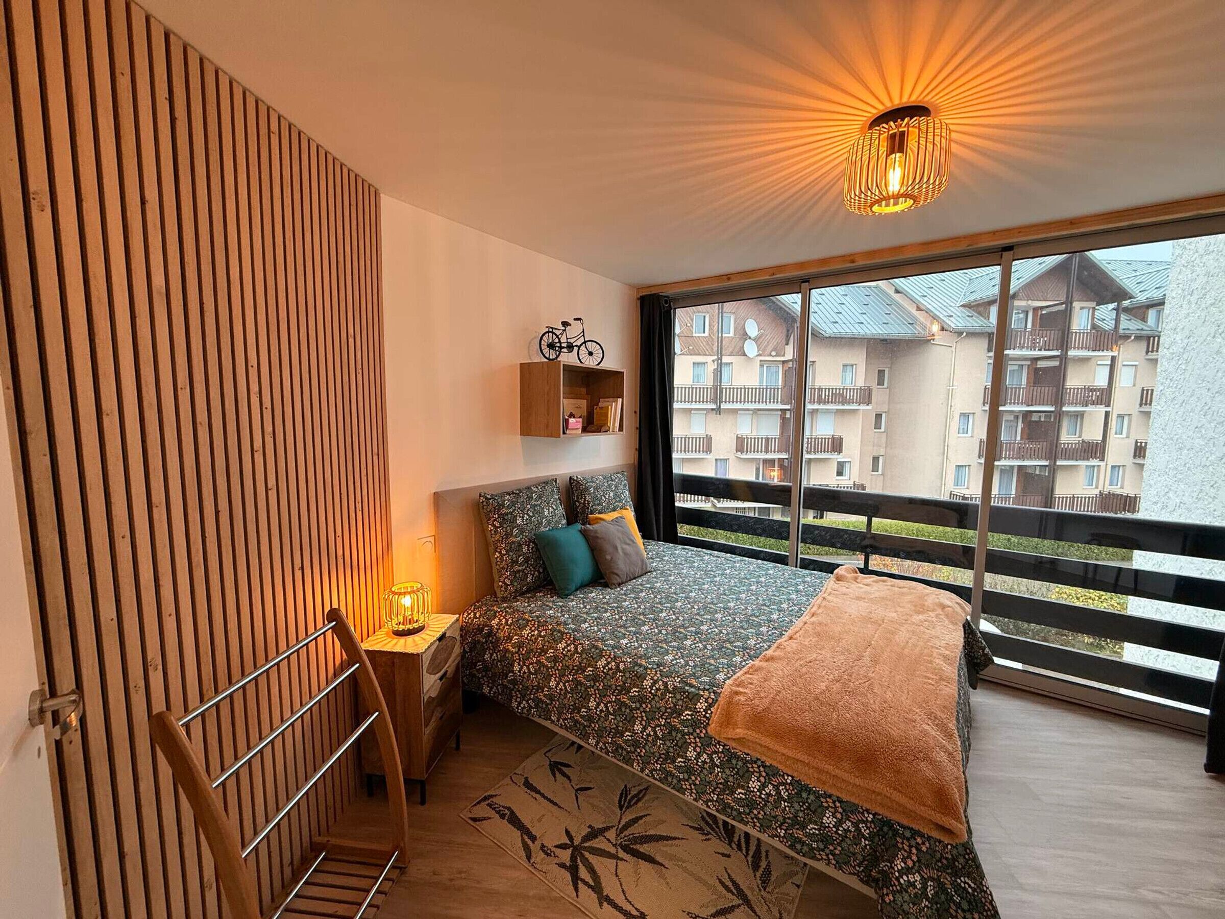A bedroom with a bed, a nightstand, a chair, a window with a view of buildings, and a ceiling light.