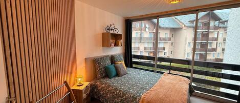 A bedroom with a bed, a nightstand, a chair, a window with a view of buildings, and a ceiling light.