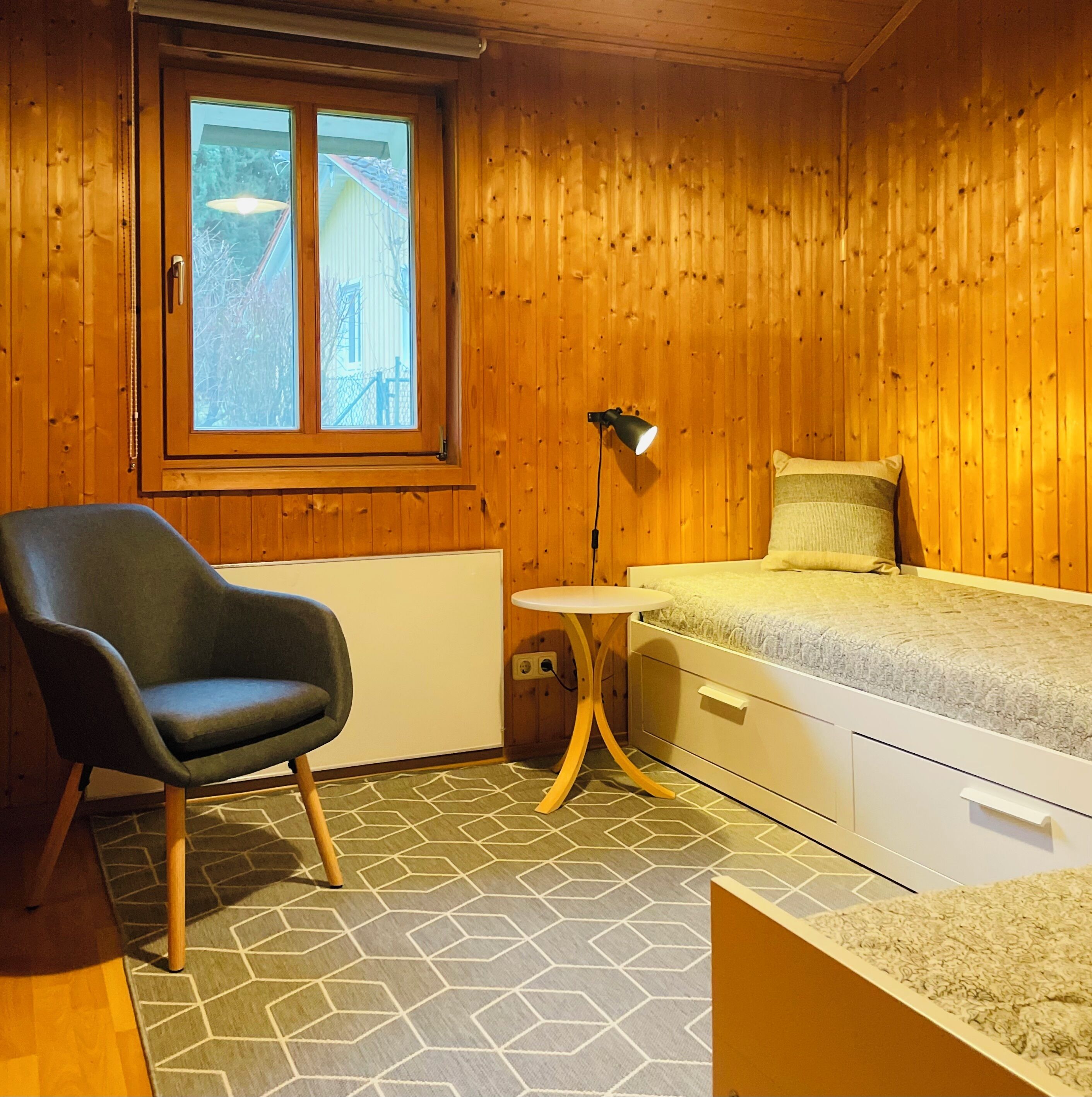 A small, cozy room with wooden walls, a single bed, a chair, and a small table.